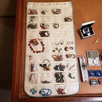 Hanging jewelry organizer filled with multiple costume jewelry items including bracelets, necklaces, brooches, and pins, displayed on a wooden table with men's watches visible to side.