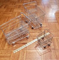 Three metal miniature grocery shopping carts of varying sizes placed on wooden floor alongside a measuring ruler to show scale.