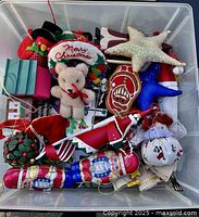 Top view of a plastic bin showing plush and textile Christmas ornaments including a teddy bear, starfish ornament, nutcracker, and other festive shapes.