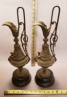 Front view of the pair of antique metal ewers showing full height alongside measuring sticks to highlight size and details of the ornate embossed metalwork and wooden bases.