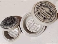 Top view of two small cream ceramic apothecary boxes with black printed labels on their lids; one labeled 'Cherry Tooth Paste Harrods Stores Ltd.' and the other 'Coty Orange Blossom Cologne Edinburgh'. Both lids are removed to show the printed information inside the boxes.