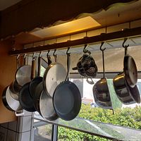 Wooden pot rack with 10 metal hooks hanging various pans and colanders by a window.