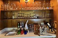 Wide view of kitchen bar shelf showing multiple rows of vintage and modern stemware including wine glasses, brandy and shot glasses, and cocktail accessories on countertop including cans, shakers, book, and beer steins.