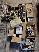 Photo of multiple trays and boxes organized with assorted screws, nails, washers, and various hand tools in cardboard boxes and plastic bins on a concrete floor.