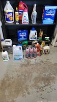 Wide shot of assorted cleaning and car care products including cleaning liquids, oils, spray bottles, and dishwasher detergent boxes arranged on and in front of a black shelf, showing product labels and quantities.