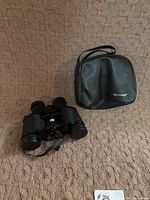 Binoculars and Tasco-branded carrying case resting on a textured beige surface.