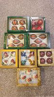 Top view of six boxed sets showing plaid and red ornaments, floral and fruit hand-decorated glass ornaments, and colorful bird ornaments.