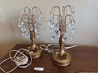 Pair of gold-tone metal table lamps with multiple hanging clear crystal pendants on curved arms, sitting on a wooden surface with white electrical cords visible.