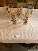 Five clear glasses with colorful Disney character prints including Winnie the Pooh, Tigger, and a character car scene. Glasses arranged on light pink towel with a note card showing lot number.