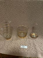 Three clear glass pieces including a jug, a bowl, and a vase with gold trim, displayed on a beige textured fabric.