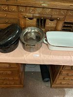 View of all three cooking items set on a table near a wooden cabinet, showing black speckled roasting pan with lid, stainless steel stockpot with glass lid and white enamel roasting pan.