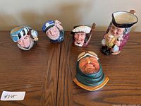 Five ceramic items on wooden table; four Royal Doulton character mugs and one bust figurine of Arctic Explorer.