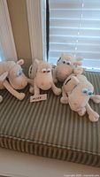 Four Serta sheep plush toys arranged on a striped cushion, showing their size, shape, and colors from front and side angles.