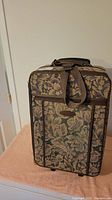 Front view of the vintage suitcase showing floral fabric, brown straps, side pockets, and handle on top.