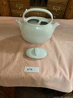 Full view of the white ceramic teapot and lid resting on a cloth background with label.