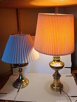 Two antique brass table lamps with pleated shades, one lit and one off, on white backdrop