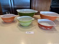 Five vintage Pyrex bowls arranged on a kitchen counter, showing varying sizes and patterns including orange with wheat design and green with floral design.
