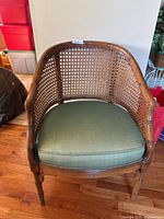 Front angled view showing wicker back, wooden frame, and green seat cushion on hardwood floor.