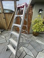 Full view of the 6-foot aluminum step ladder standing outside on a stone patio, showing the front steps and top platform with brush and tool holders.