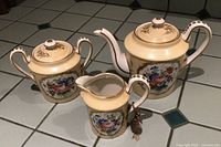 Full shot of teapot, sugar bowl, and creamer on tile surface showing floral painted panels and gold trim