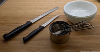 Items arranged on wooden surface: 4 white ceramic bowls stacked, 2 knives, set of 4 measuring cups, 3 wire whisks