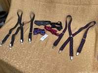 Full lot of suspenders and bowties arranged on a beige textured surface. Two pairs of suspenders on right and left, four bowties in center.