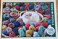 Front of the 1000 piece jigsaw puzzle box showing a white cat in a basket surrounded by colorful yarn balls by Cobble Hill.
