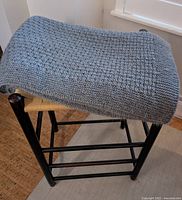 Pale blue-green hand-knitted lap blanket folded on wooden stool showing knit texture and color.
