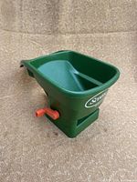 Side view of green Scott branded handheld seed spreader with red adjustment lever.