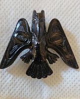 Front view of black Indigenous-style raven brooch with detailed engraved wing and tail feather patterns displayed against a white textured background.