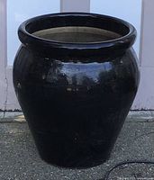 Full view of the large glossy black ceramic plant pot showing its shape and size against an outdoor fence.