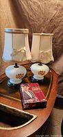 Photo showing two white glass lamps with floral designs on a wooden table, each topped with a cream fabric lampshade with gold trim and bows, along with the boxed DVD set of Agatha Christie's Miss Marple.