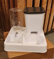 White plastic pet food and water dispenser with a clear water bottle and black lid food compartment