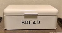 Front closed view of cream coloured enamelled metal bread box with 'BREAD' printed on front in black.