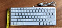 Top view of white Apple keyboard showing full key layout and attached USB cable, tape measure shows approx 11 cm height.