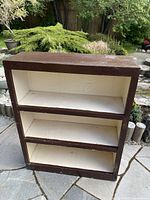 Front view of the painted wooden shelf unit showing three shelves with white interior and dark brown exterior, placed outdoors on stone patio.