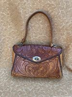 Front view of vintage handtooled leather handbag showing floral and leaf pattern on flap and body, braided edge, brown leather strap and silver-tone twist lock.