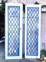 Full view of the two vintage French doors leaning side by side showing diamond leaded glass and white wooden frames with brass knobs.