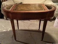 Front view of the oval leather topped plant table showing the wood frame, leather surface with gold embossing, and the metal inserts on both ends.
