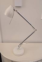 White adjustable desk lamp on a white surface against a plain wall, showing the full lamp with on/off switch on cord.
