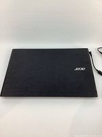 Closed black Acer Aspire laptop with power cord visible, top view showing Acer logo and textured plastic surface with minor wear.