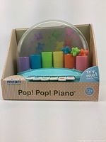Front view of new Mirari Pop! Pop! Piano baby toy in its original packaging, showing colorful piano keys and butterfly pop-up cylinders under clear dome.
