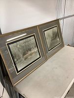 Two framed vintage sketches resting against wall on table, showing size and framing style.