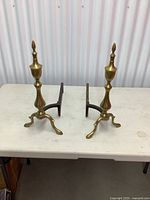 Pair of vintage brass fireplace andirons on a white table, showing front and side views with turned urn-shaped columns and finial tops, three-legged bases.