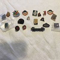 Photo of assortment of collectible pins on white cloth surface showing various designs including cartoon faces, logos, and multicolored symbols.