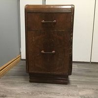 Frontal view of the antique wooden bedside table showing upper drawer and lower cabinet door with wooden handles, standing on a stepped base on a wooden floor.