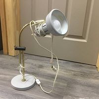 Single photo showing a grey metal desk lamp with brass adjustable arm and clamp, round grey base, and attached power cord with inline switch.