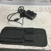 Photo showing Garmin Nuvi 250EB GPS device with cable and a black sun visor organizer with several pockets on a white patterned fabric surface.
