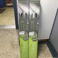 Two new Gutterware gutter covers in original packaging, standing upright on floor.