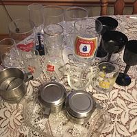 Miscellaneous glassware displayed on a lace tablecloth including beer glasses, a Coca-Cola glass with metal strainer, black goblets, whiskey branded glass, two glass containers with metal lids, all on a metal tray.
